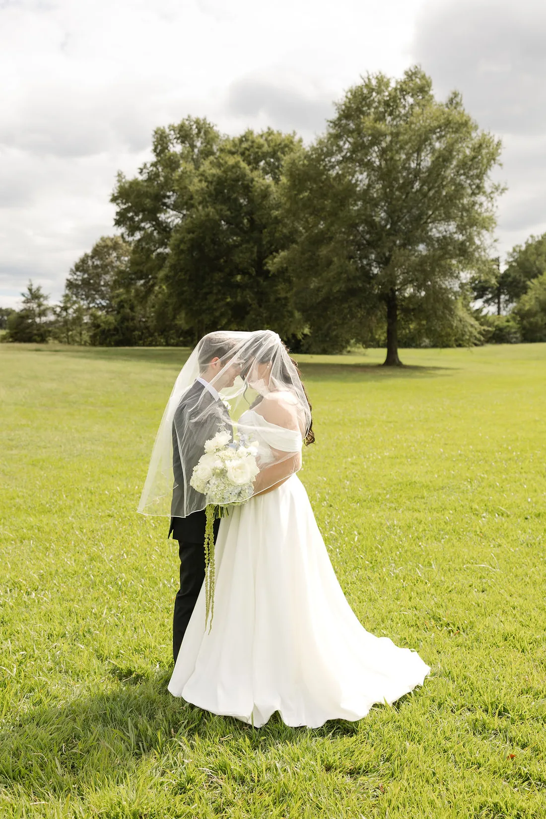 Jennifer and Hayden sharing a kiss under her veil in the field at Gateley Hill Gardens